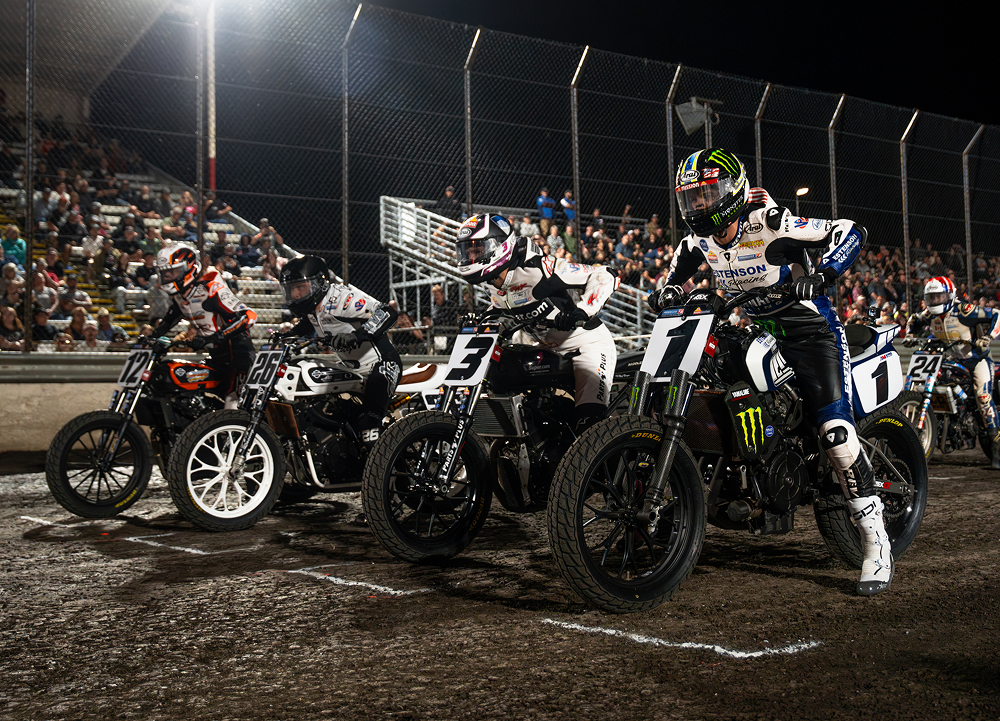 Flat track motorcycle racers launch from the starting line during a nighttime race as spectators fill the grandstands behind the dirt track