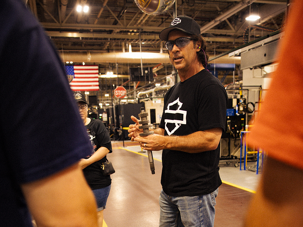 Factory guide holding a gear shaft with machinery and an American flag in the background