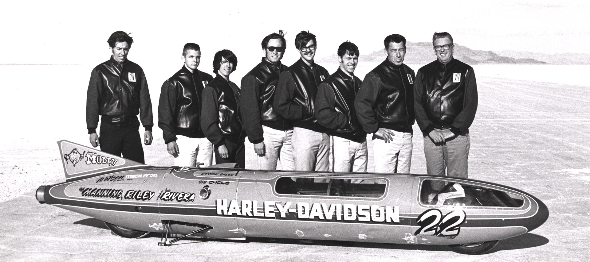 Eight men in black Harley jackets stand behind the Sportster streamliner in the desert.