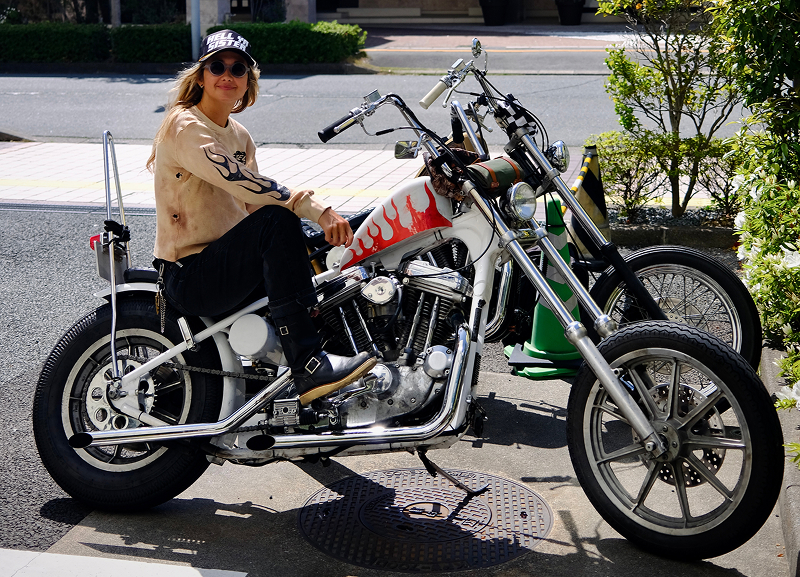 Becky Goebel seated on a custom chopper with flame-painted tank parked curbside on a sunny street with greenery nearby