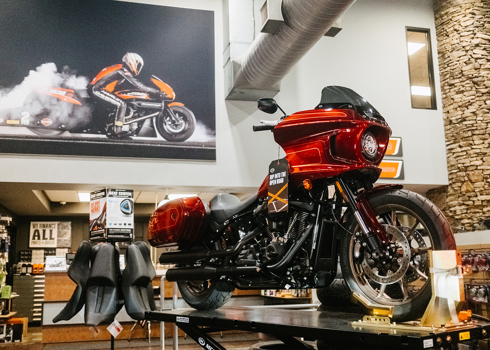A red Lowrider ST on a platform in the showroom of Moonshine Harley-Davidson with accessories and a large photo of a drag bike burning out in the background.