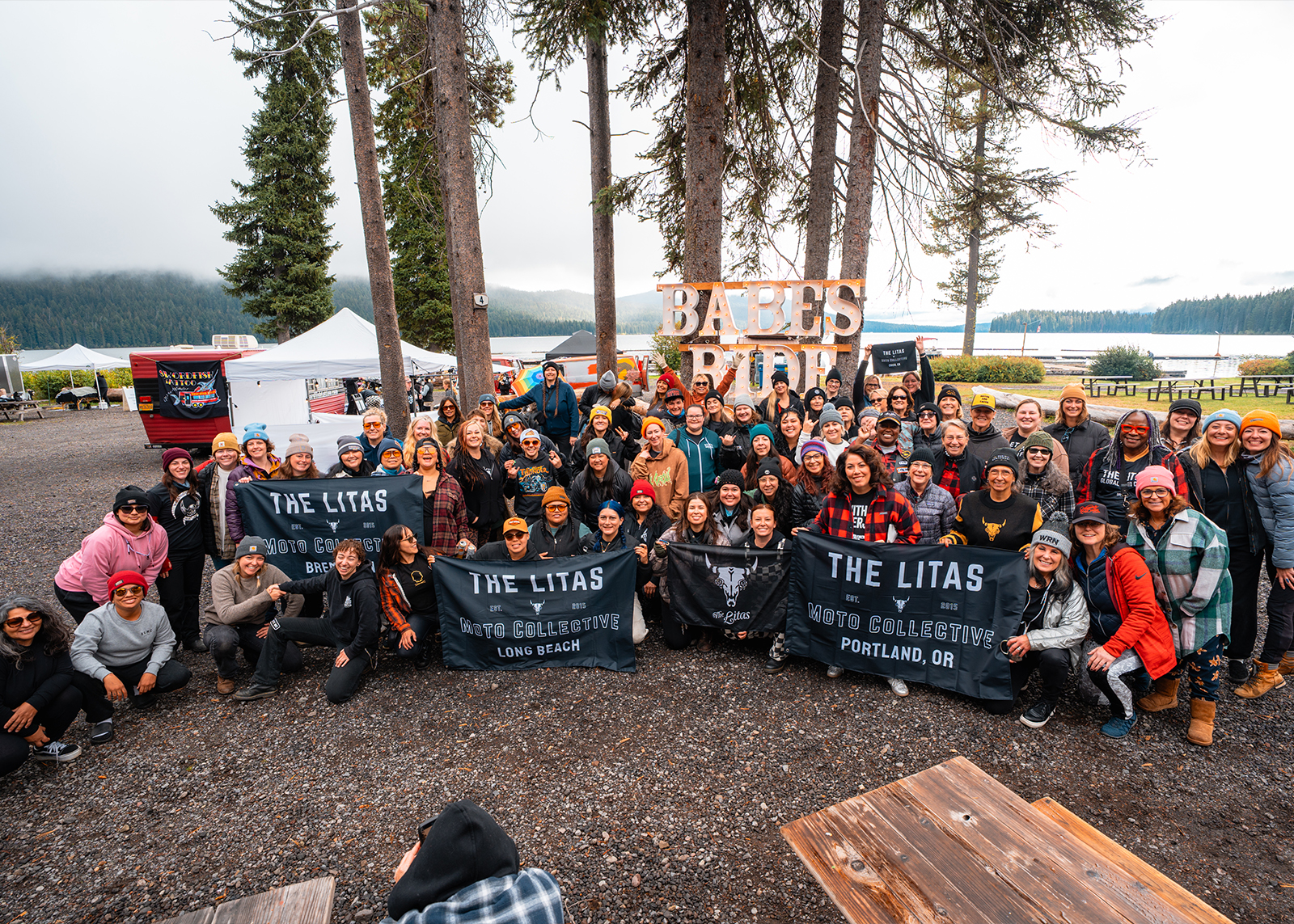 A large group of women riders pose lakeside with The Litas Moto Collective banners at the Babes Ride Out event.