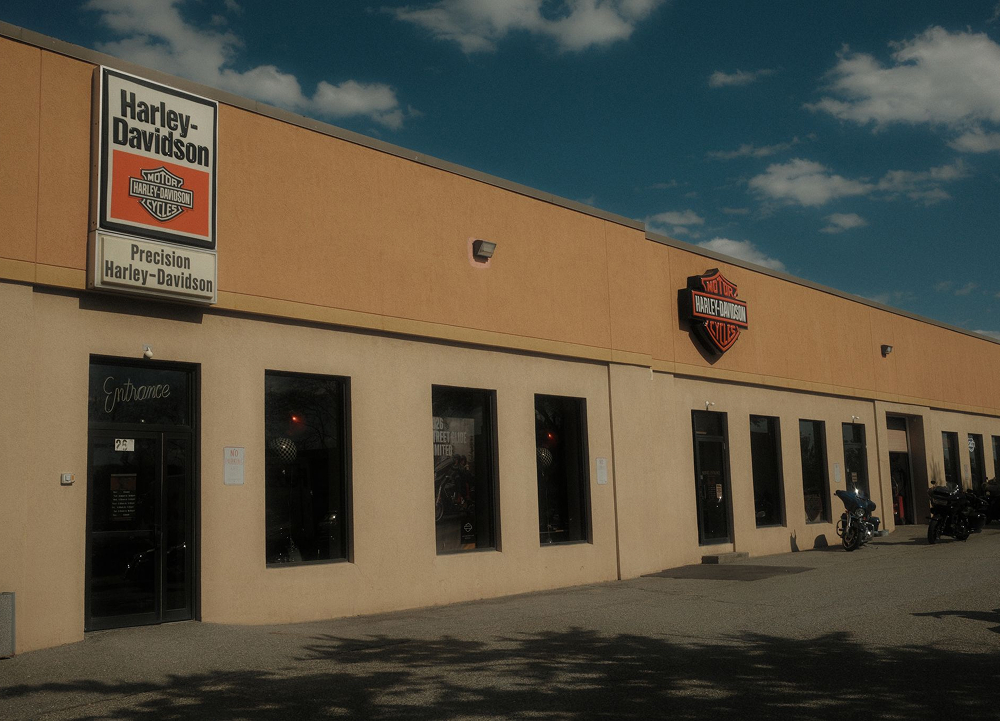 Precision H-D dealership exterior with tan facade, signage, windows, and motorcycles parked outside under blue sky
