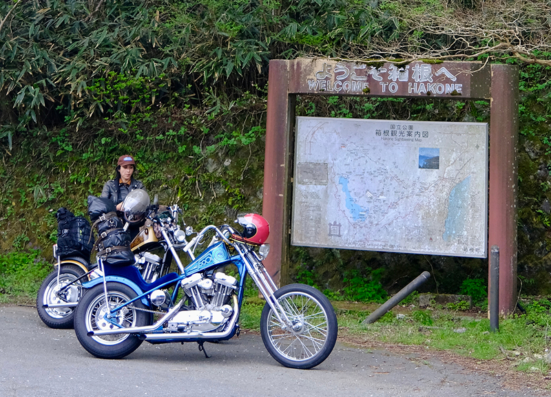 Two custom choppers parked beside a roadside sign reading welcome to hakone with Adri Law standing nearby
