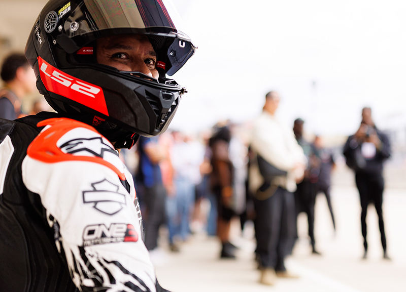 Close-up of a helmeted motorcycle racer standing in the paddock with blurred figures and garages behind