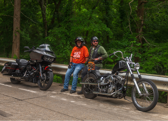 Payton and Barry Broussard standing roadside with two motorcycles parked on a bridge, backed by lush forest scenery