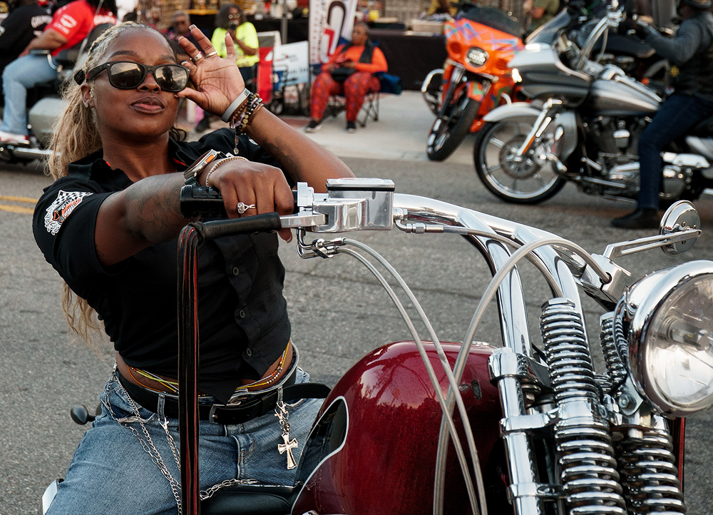 Rider gripping tall handlebars on a custom motorcycle during a crowded street gathering with bikes nearby