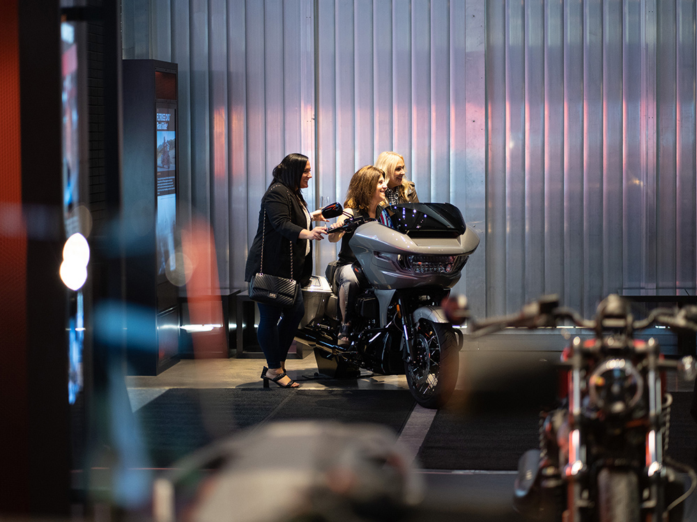 Visitors sitting on motorcycle while another person stands nearby in museum exhibit