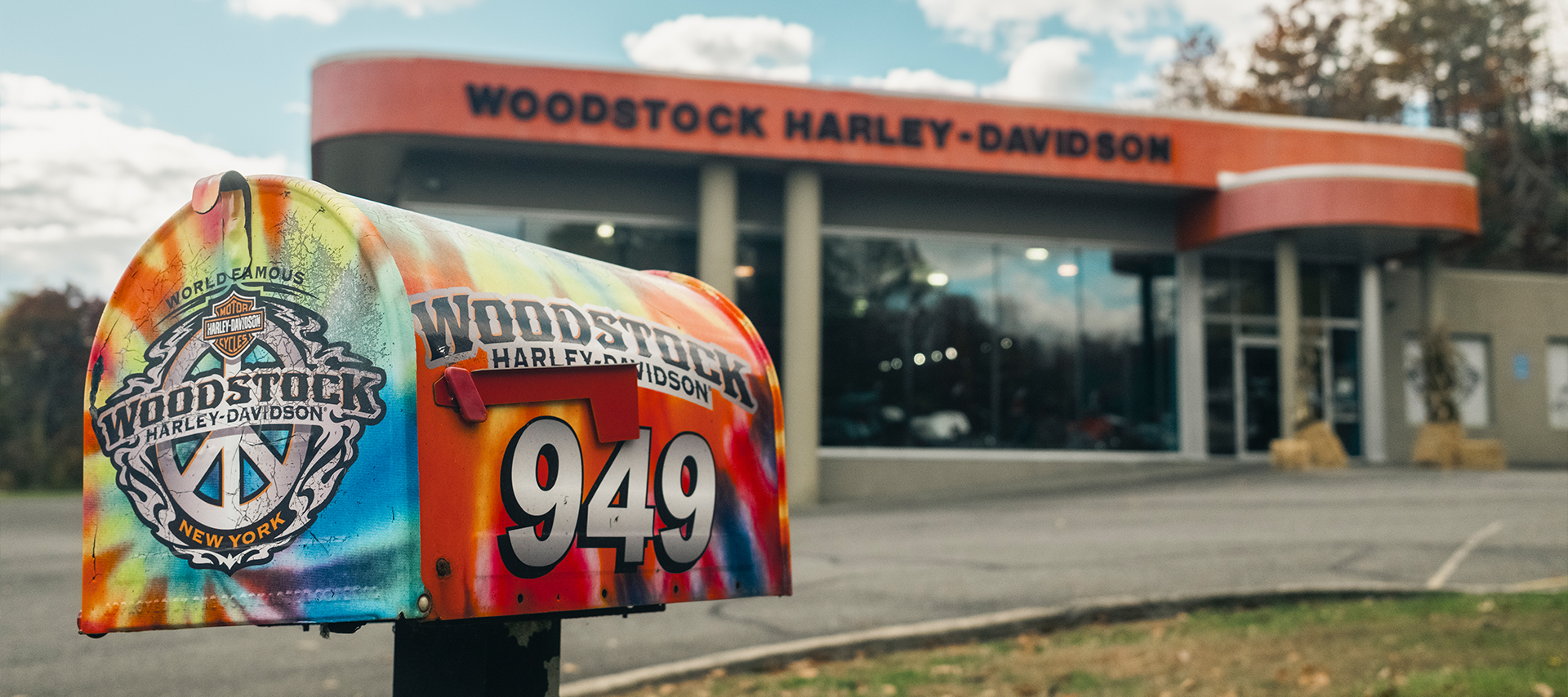 Colorful tie-dye style Woodstock Harley-Davidson mailbox with peace-sign logo in front of the dealership building.