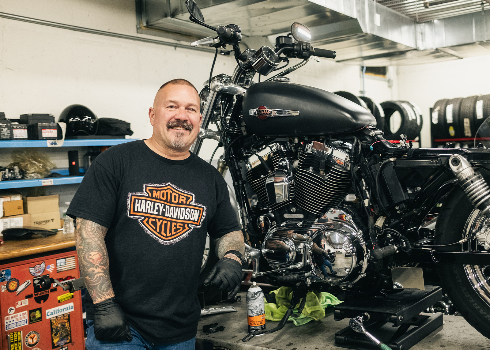 A mechanic poses and smiles standing next to a motorcycle on a lift at Harley-Davidson of Nassau County.