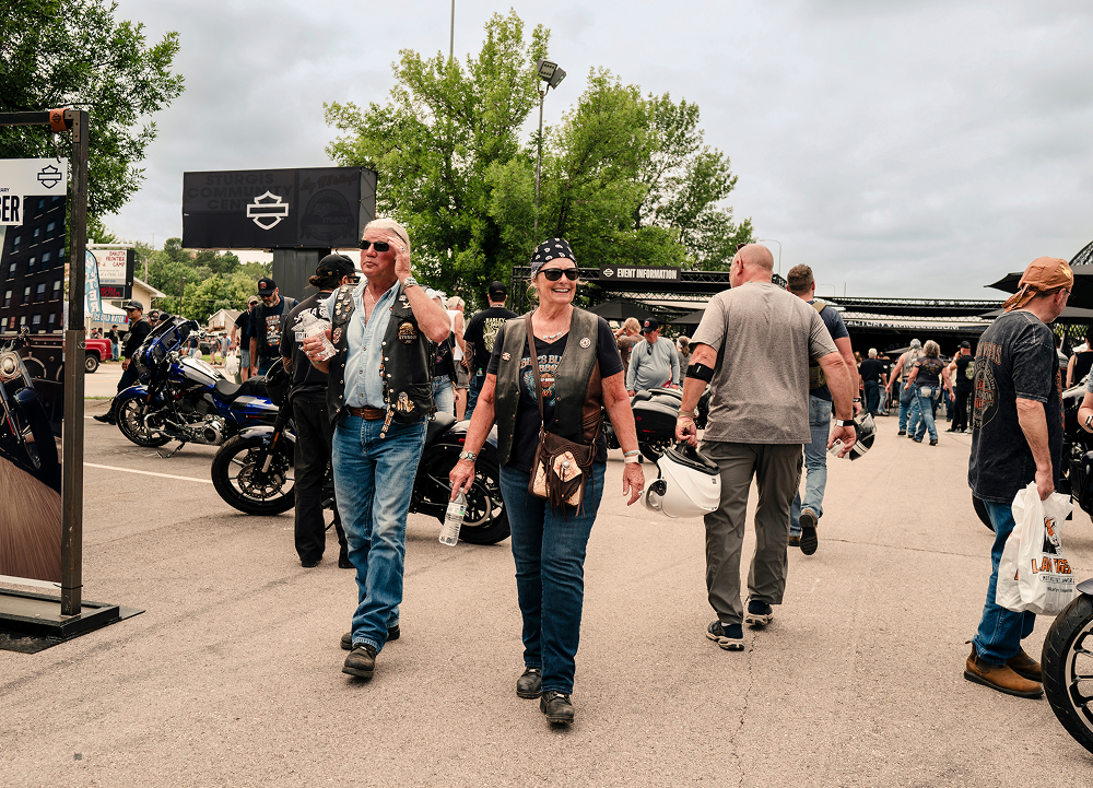 People walking through busy H-D rally street with motorcycles parked and vendor booths in background.