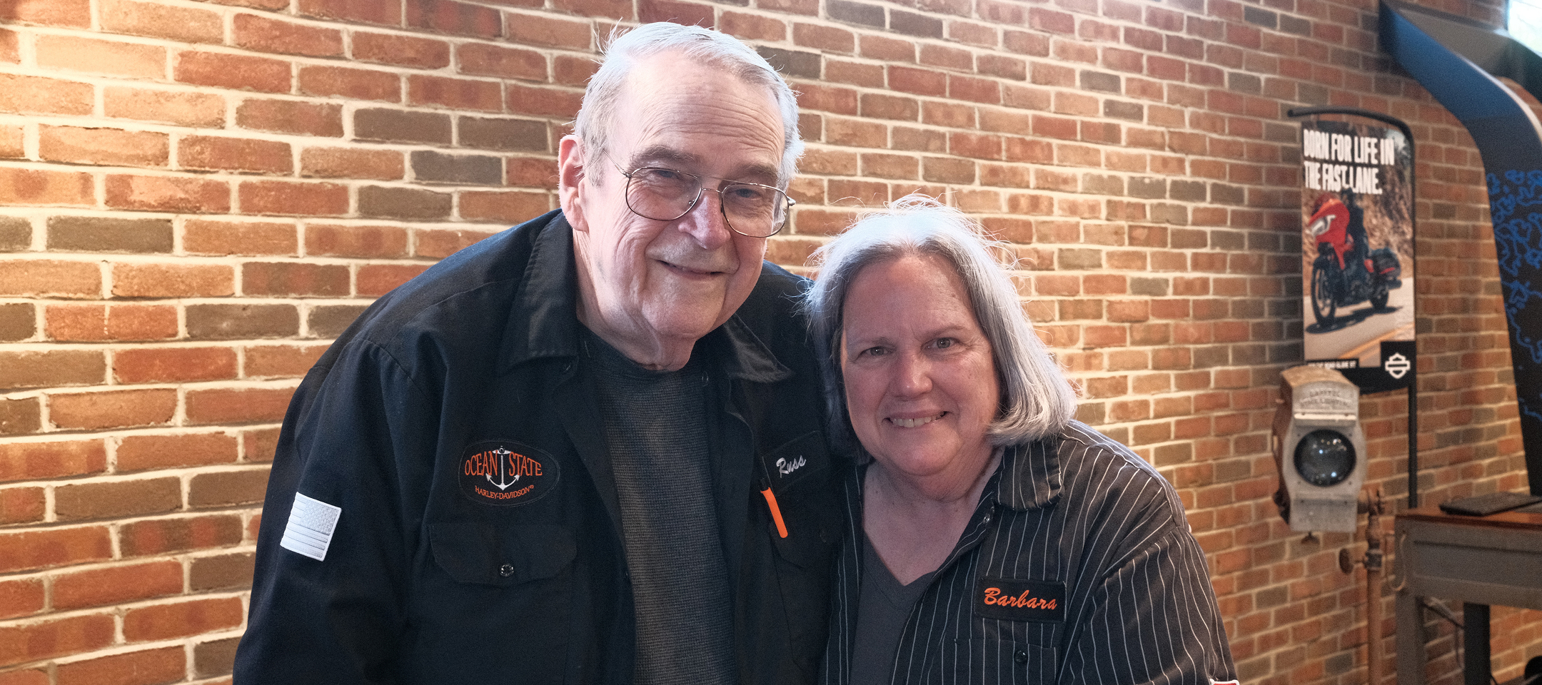 Founder Russ Hampton poses with long time employee Barbara in front of a brick wall inside Ocean State Harley-Davidson