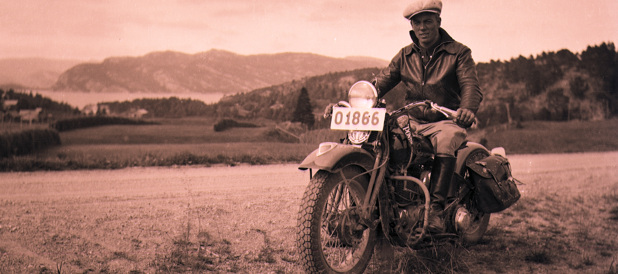 Vintage photo of Gustaf Stenmark seated on a classic motorcycle with saddlebags, parked overlooking hills and water in the distance