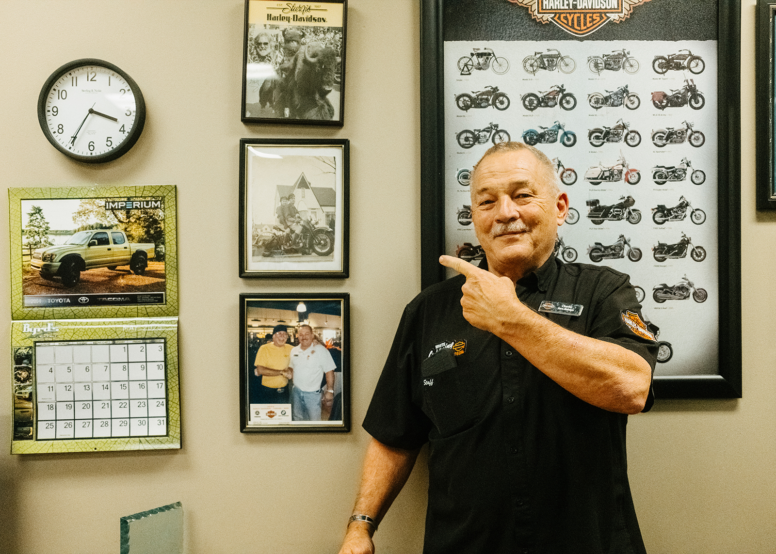 A White Lightning Harley-Davidson employee points towards a wall with various framed posters and photos.