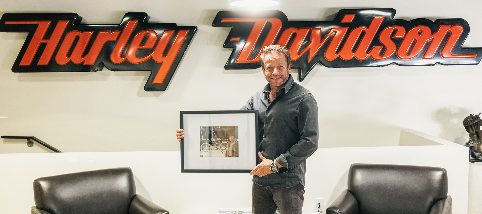 Harley-Davidson of Nassau County Owner and Dealer Principal Larry Altholtz poses holding a framed photo in front of a large Harley-Davidson sign hanging on the wall.