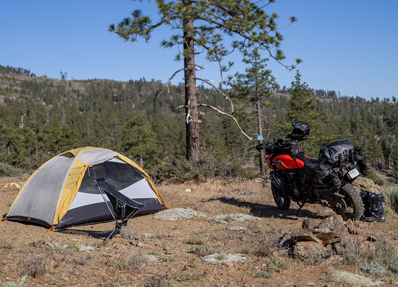 Red H-D Pan America motorcycle parked beside tent at remote campsite with chair, trees, and hillside landscape under clear sky
