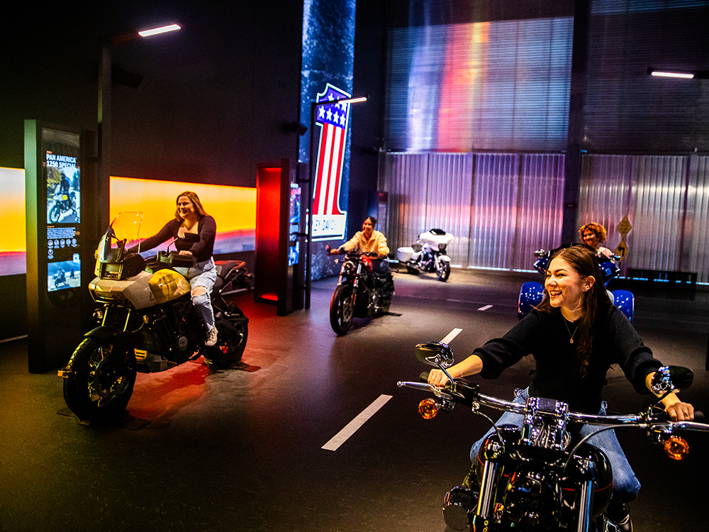 Visitors riding motorcycles inside interactive museum exhibit with illuminated backdrop