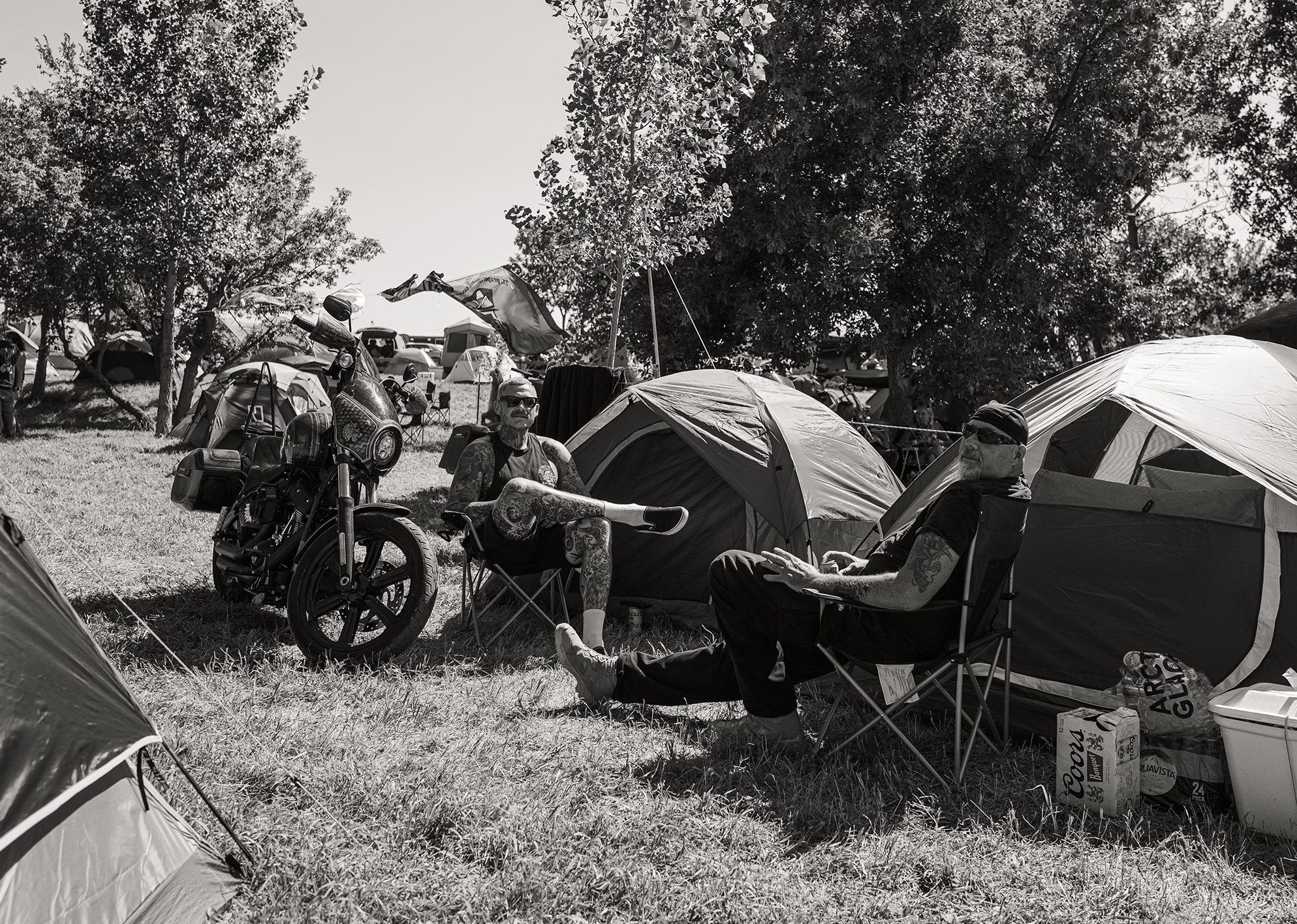 Two bikers relax in camp chairs beside tents and a motorcycle at a rally campsite.