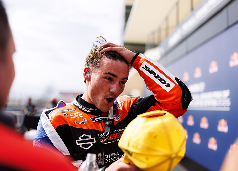 Motorcycle racer in branded leathers stands trackside pushing his hair back while holding a yellow hat in the other hand