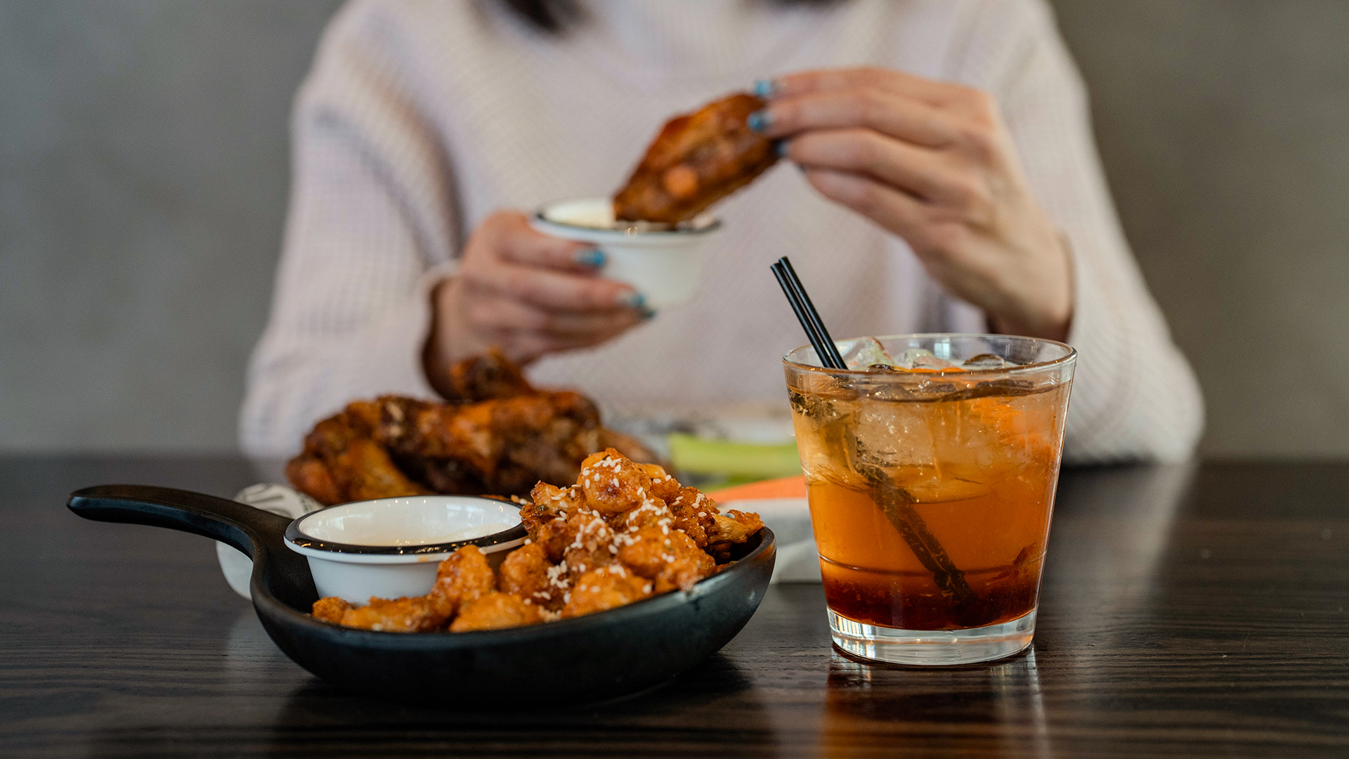 Person dipping fried appetizer into sauce with drink on table