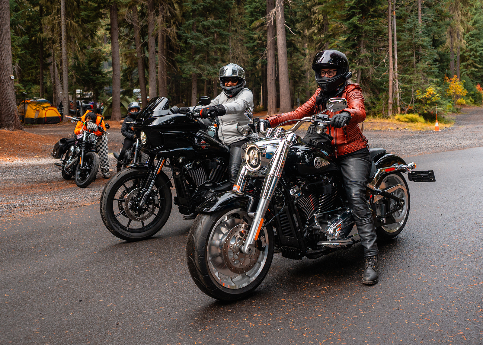 Motorcyclists in helmets and jackets prepare to ride through a wooded campground road.