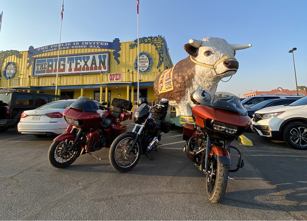 Three motorcycles parked in front of The Big Texan Steak Ranch, with a large cow statue beside the entrance