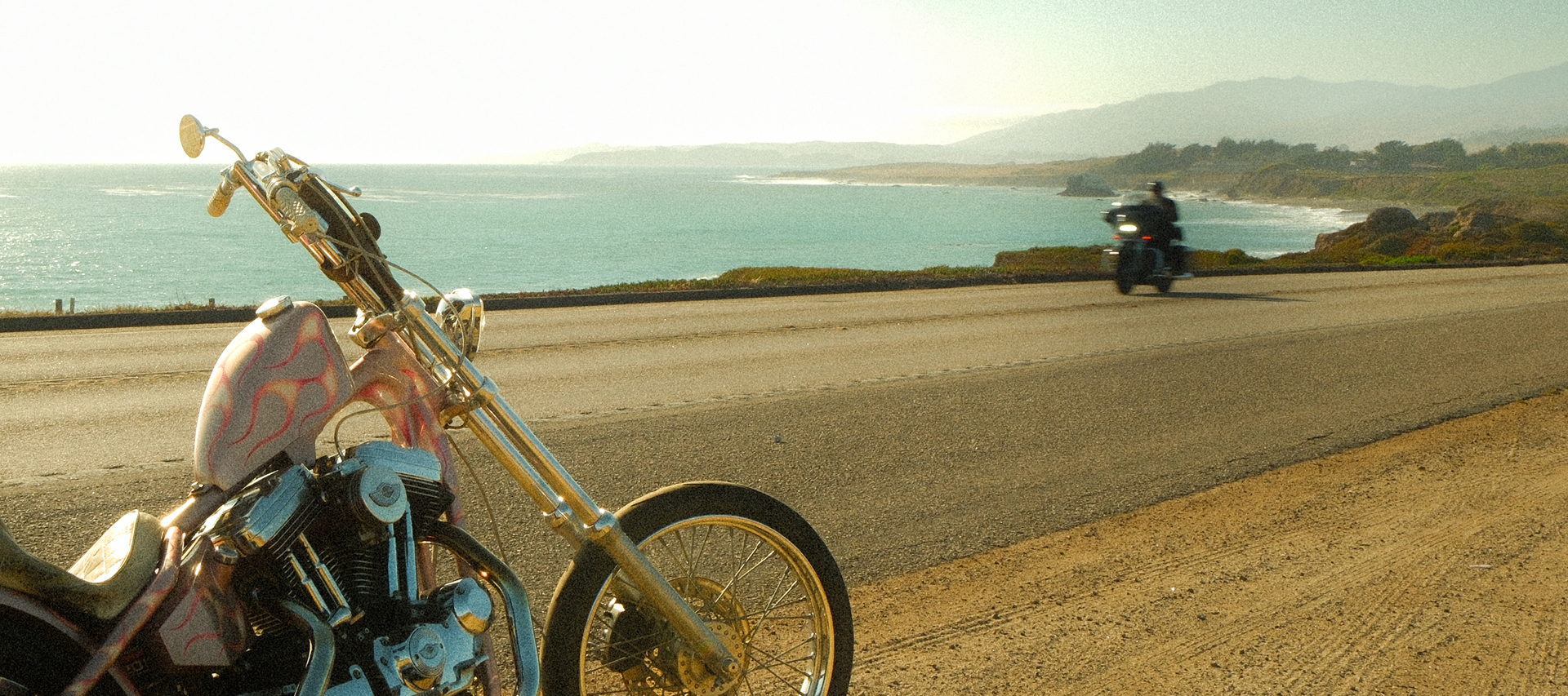 Custom chopper motorcycle parked along a coastal highway with ocean views, rugged shoreline, and passing rider in the distance