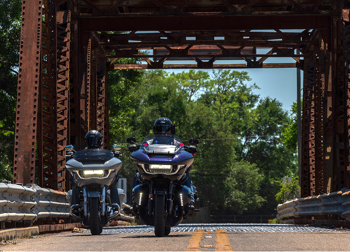Two touring motorcycles ride side by side across a narrow steel bridge, framed by rusted trusses and trees
