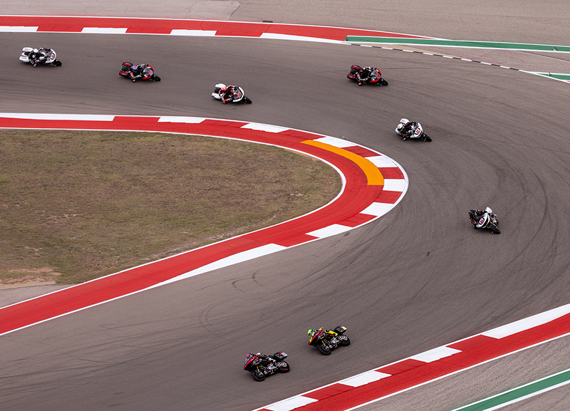 Aerial view of multiple motorcycles spread out through a sweeping racetrack turn with red and white curbing