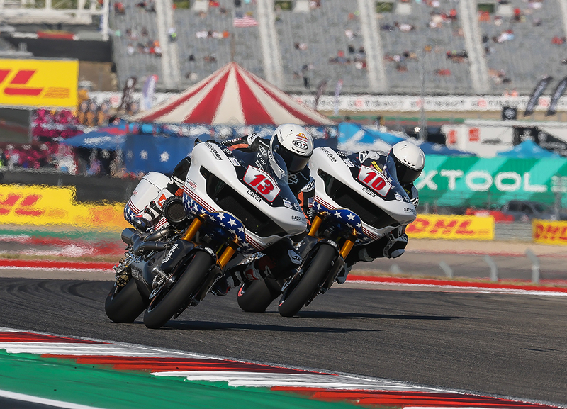 Two motorcycle racers accelerate together through a turn, tightly grouped on a professional track