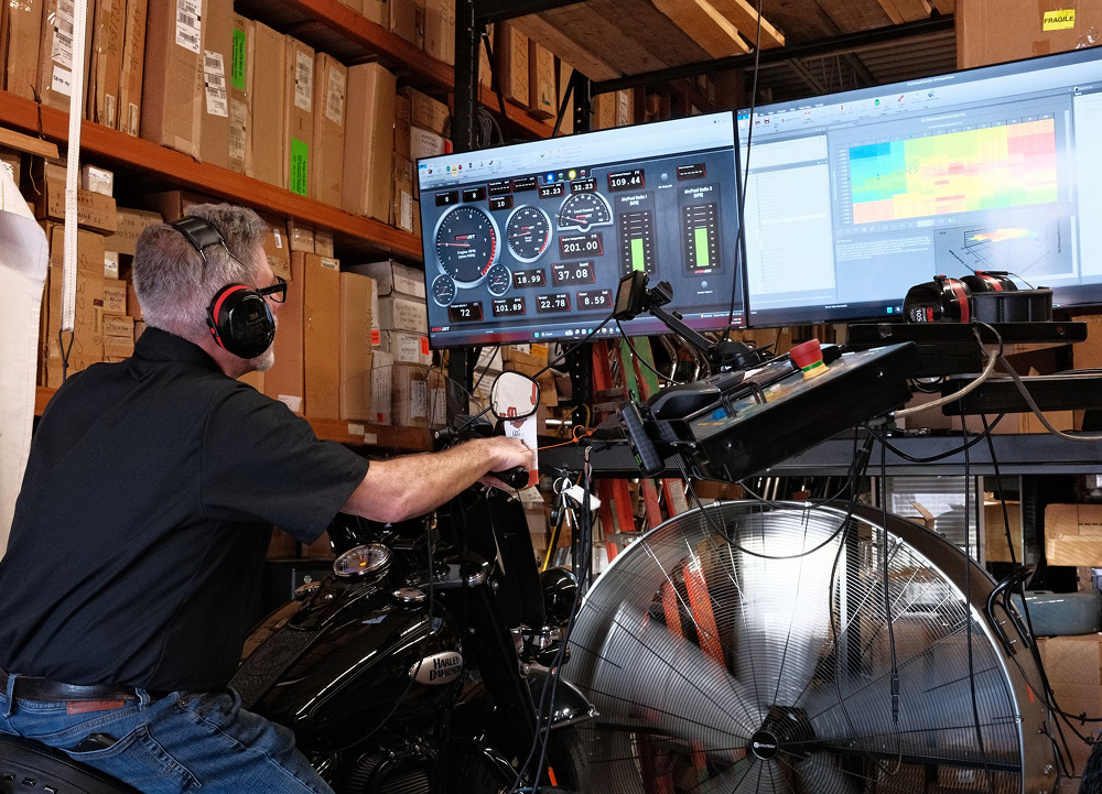 Technician tuning hd motorcycle on dyno with computer monitors displaying performance data and cooling fan nearby