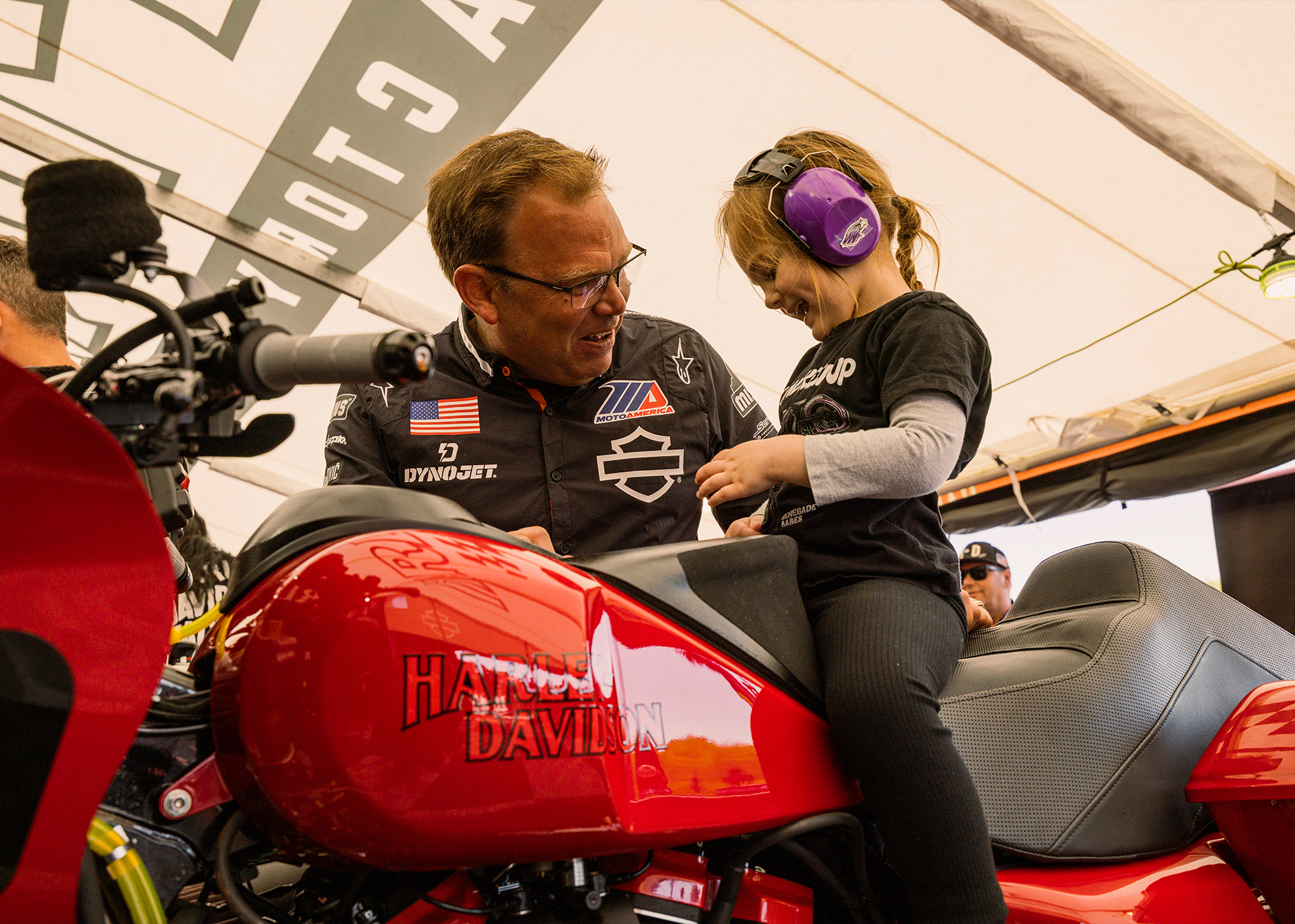 Harley-Davidson Factory Racing Lead Crew Chief Bjorn Christensen helps a child sit on a red Harley-Davidson race motorcycle in a tent.