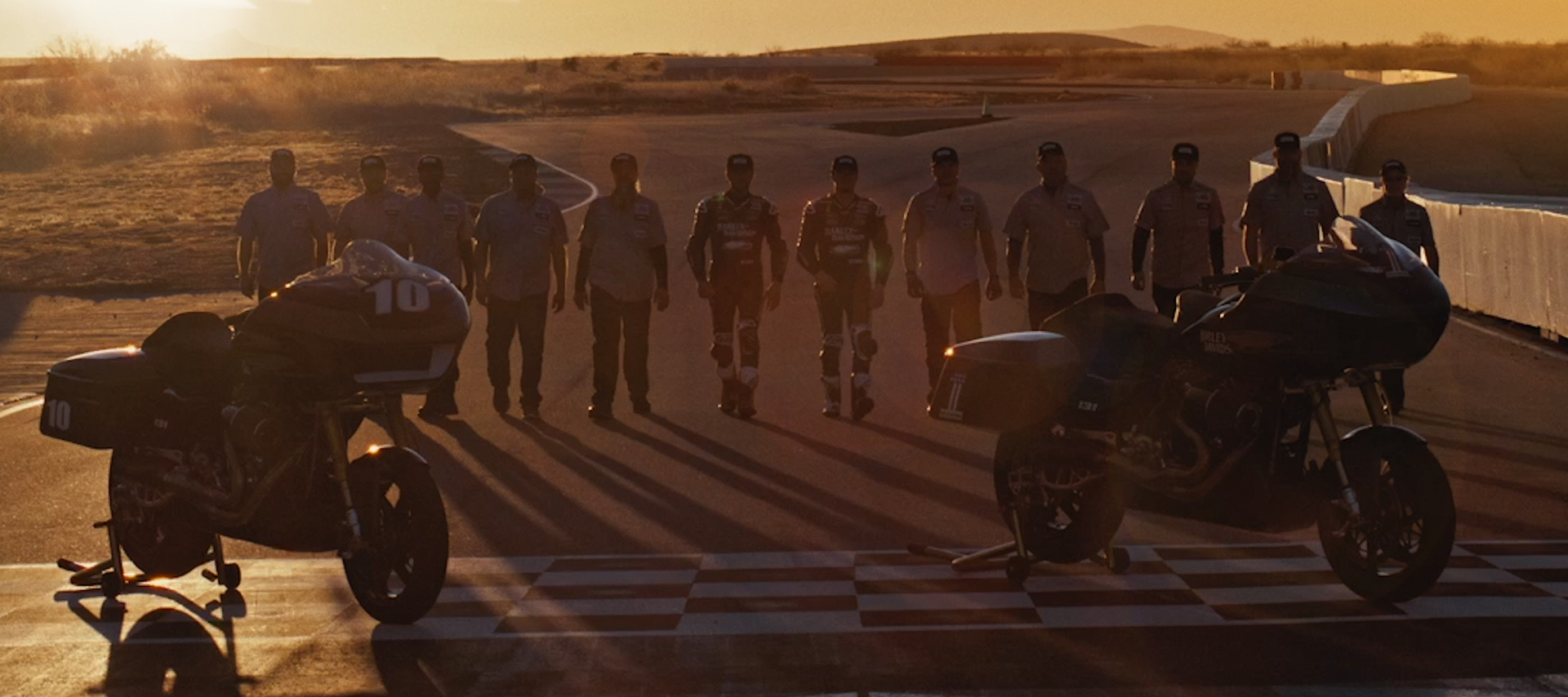 Two race bikes parked on a track with a line of people walking toward them.
