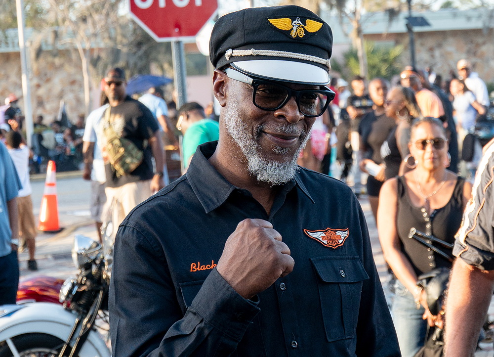 Motorcycle personality Blackula raises a fist while standing on a crowded street lined with bikes and spectators