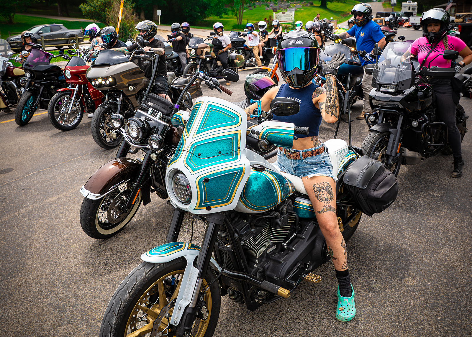 A woman in crowd of bikers parked on the road waves towards the camera.