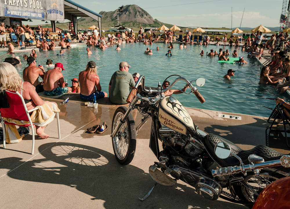 Custom H-D chopper parked beside crowded pool with riders relaxing and mountains in background.