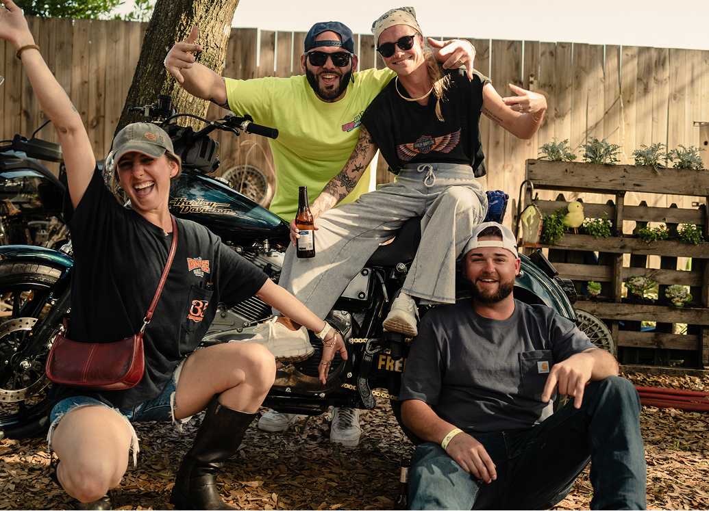 Group of friends posing with motorcycles outdoors, sitting and standing near bikes in a casual yard setting