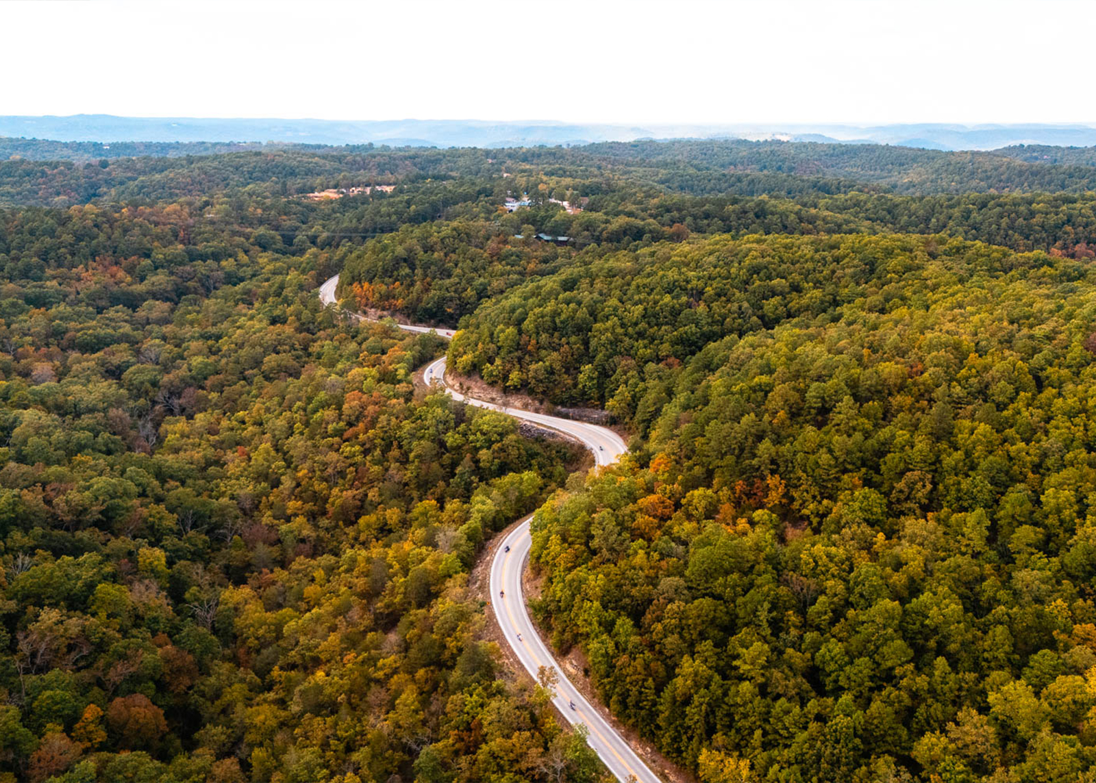 A winding two-lane road cuts through rolling hills with foliage about to change into autumn colors 