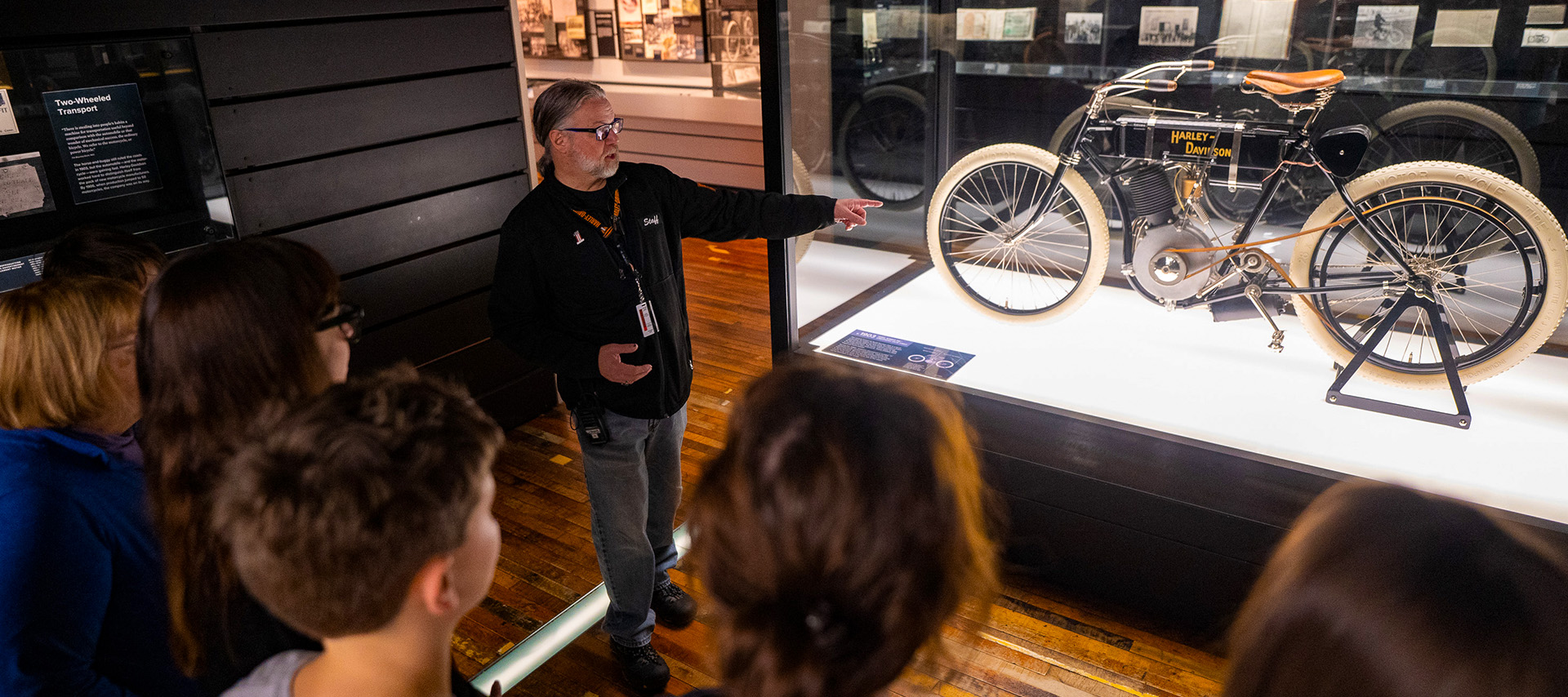 Guide pointing at vintage motorcycle in glass display while visitors watch