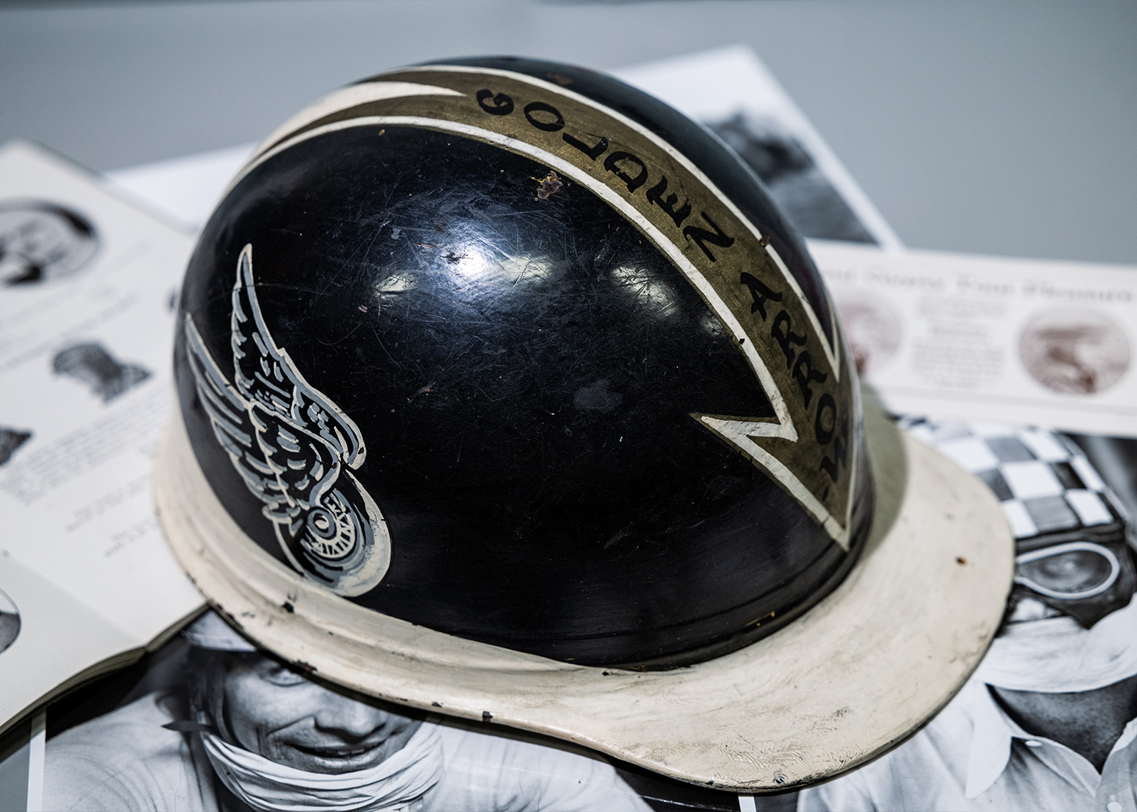 A vintage black plastic motorcycle helmet with a build in white visor, winged wheel graphic, and golden arrow pointing forward.