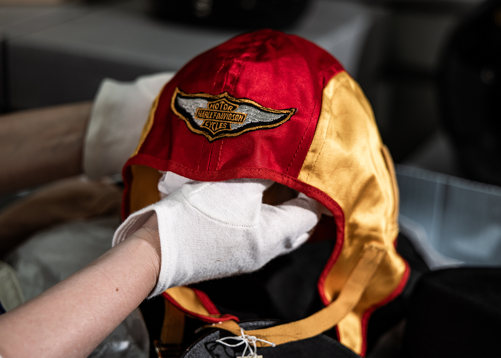 A museum archivist holds a vintage red and gold satin motorcycle helmet with winged Harley-Davidson logo on the front.
