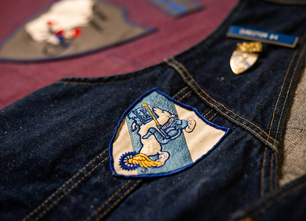 Close-up of dark denim vest fabric with a stitched patch, visible seams, metal snap, and textured embroidery