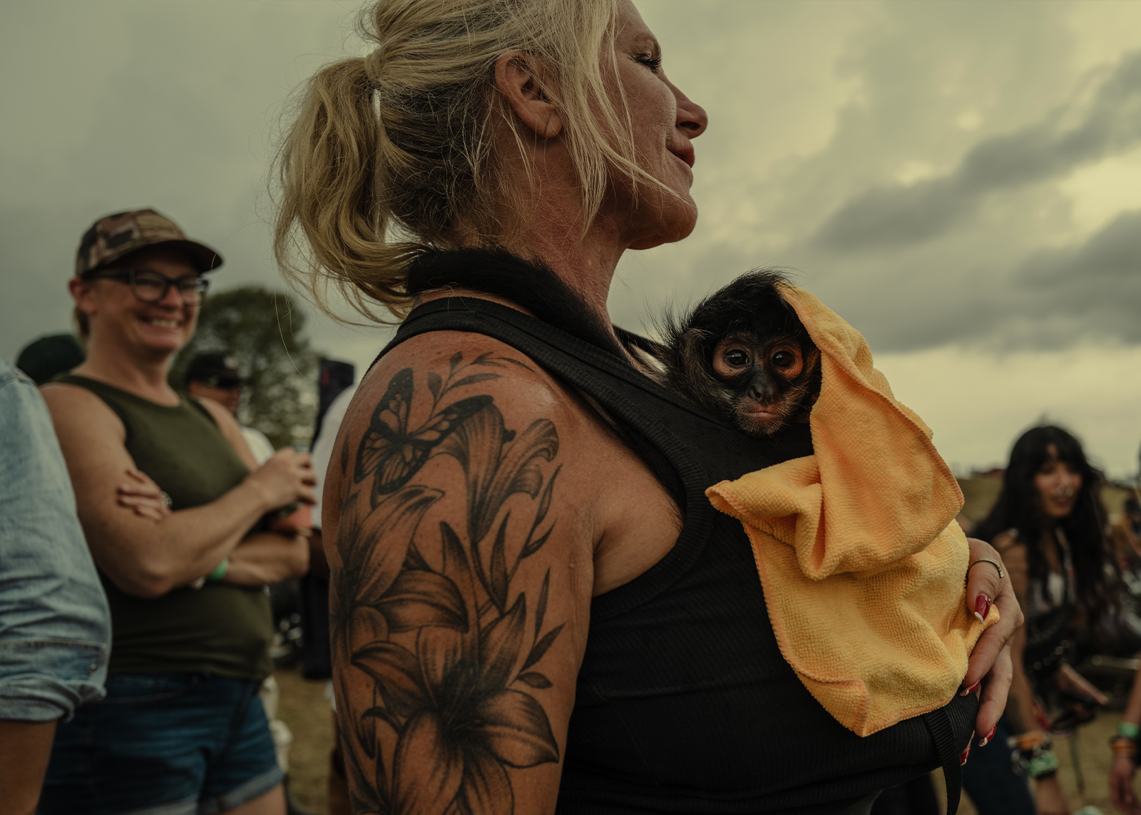 A woman with a floral tattoo holds a small monkey wrapped in a yellow cloth amid a smiling crowd.