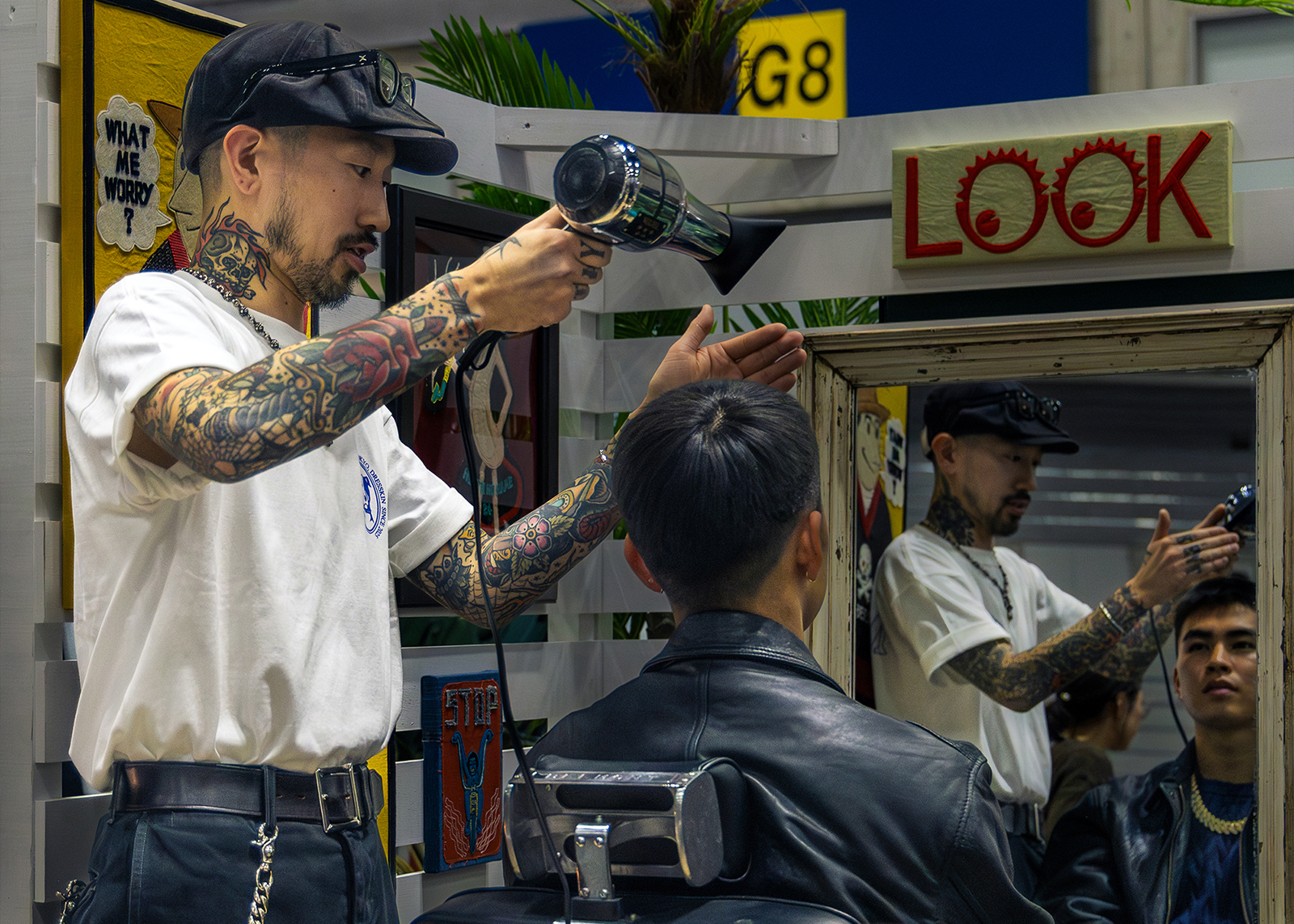 A tattooed hairstylist wearing a white T-shirt and black cap uses a hair dryer on a seated client