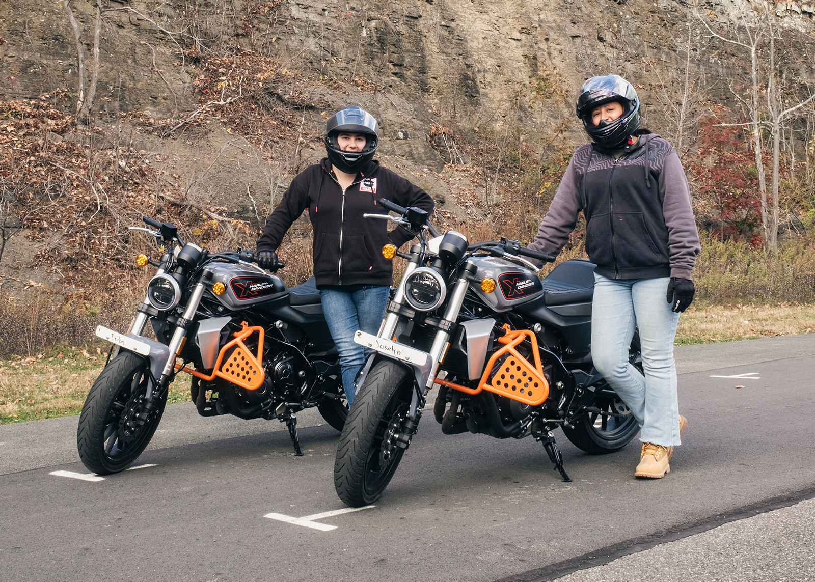 Two H-D® Riding Academy students wearing helmets pose with motorcycles in the parking lot at Woodstock Harley-Davidson.