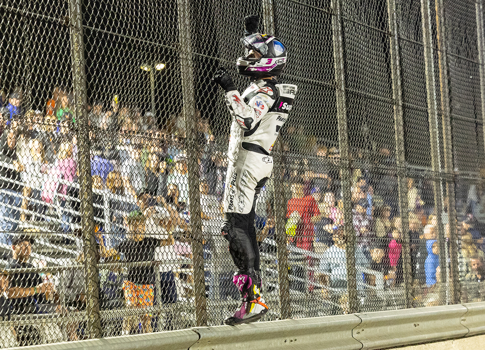 Flat track racer climbs the catch fence after a nighttime victory as fans fill the grandstands behind