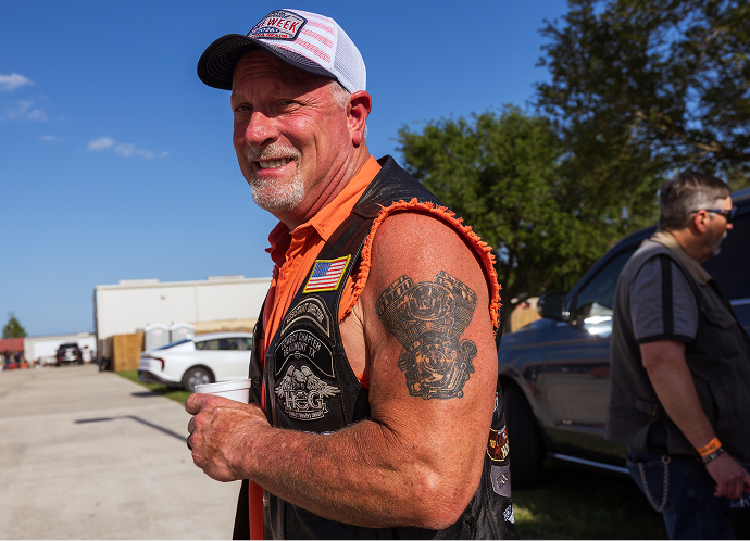 A biker wearing a sleeveless vest and cap smiles while showing his Harley-Davidson engine tattoo on his bicep