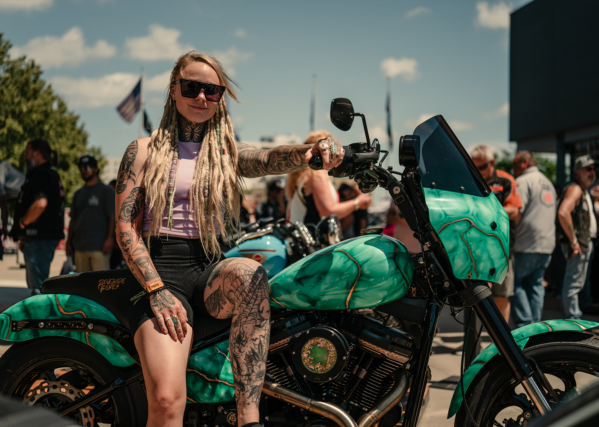 A young woman with blonde dreadlocks, tattoos, wearing sunglasses sits on her custom motorcycle
