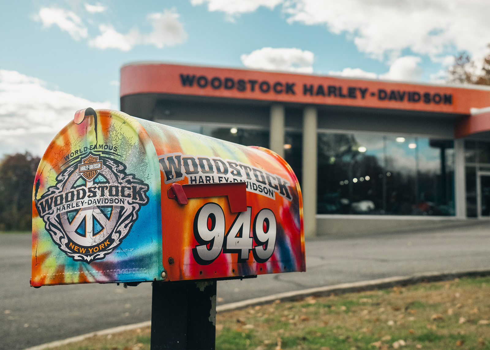 Colorful tie-dye style Woodstock Harley-Davidson mailbox with peace-sign logo in front of the dealership building.