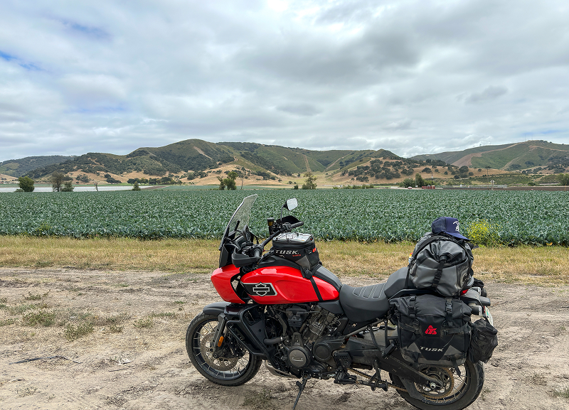 Red H-D Pan America motorcycle with full luggage parked beside farmland and rolling hills under clouds